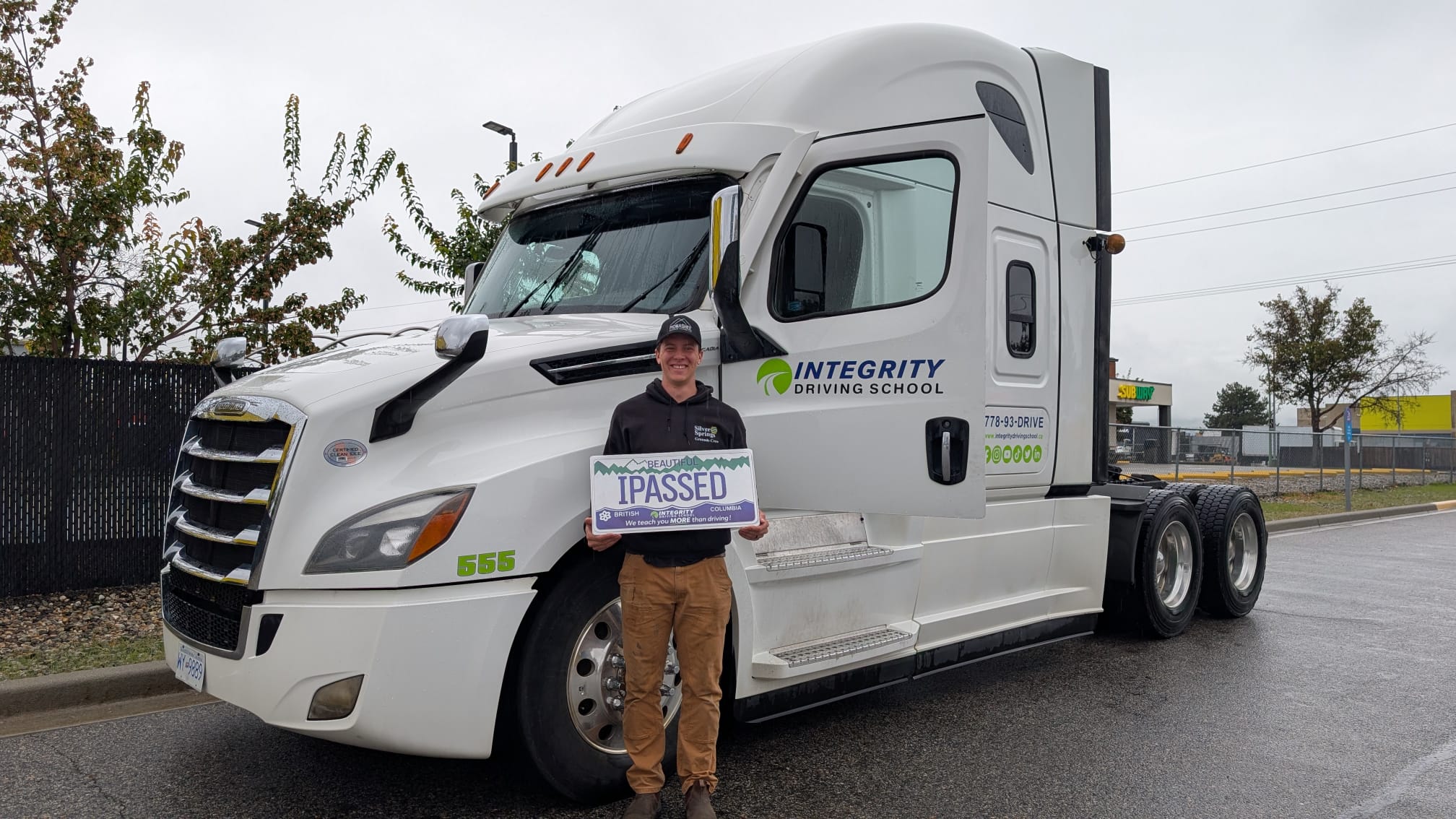 Integrity Driving School Class 3 graduate standing beside a branded Freightliner Cascadia truck