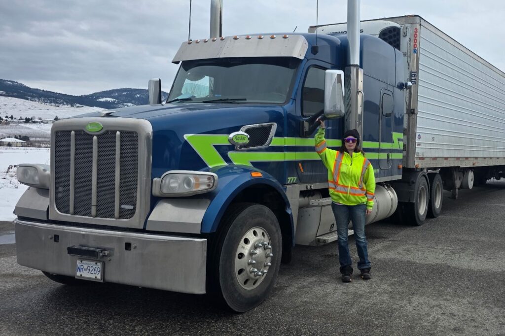 Commercial driver in high visibility gear beside a Peterbilt tractor and reefer trailer on a snowy Okanagan lot